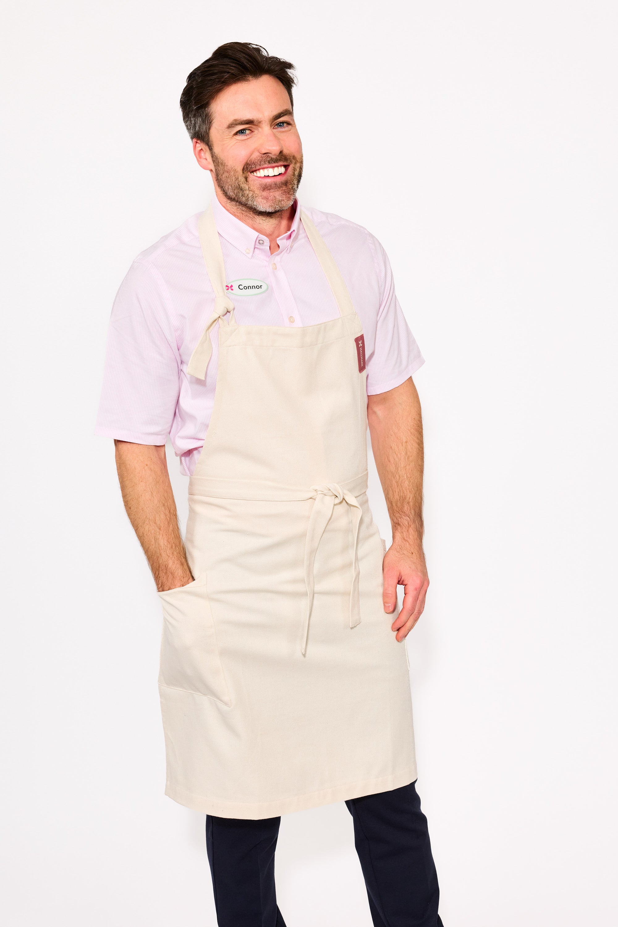 Man wearing a beige apron over a pink shirt with a visible brand logo on a white background